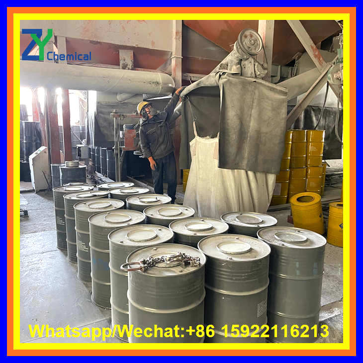 A factory worker wearing protective gear manages the industrial packaging process of calcium carbide powder into grey metal drums.
